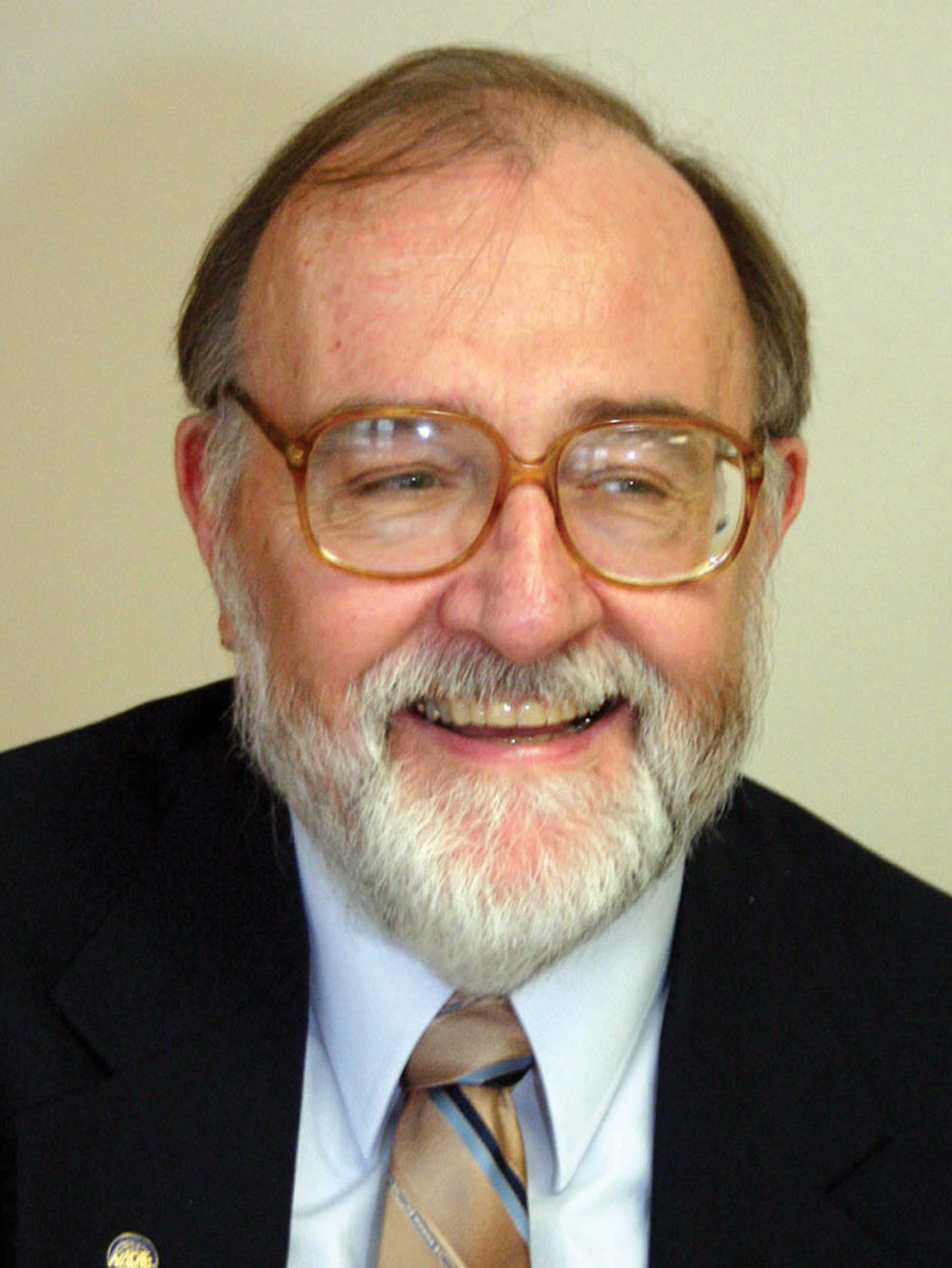 A smiling Donald L. Sweeney in tortoise shell glasses and a suit with a gold tie
