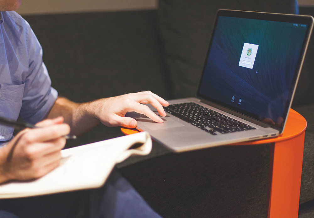 Man clicking on a laptop keyboard trackpad/touchpad area while holding a pen and notepad as he glances at his laptop screen display showing a log in password entry
