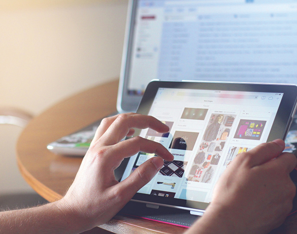 Close-up portrait photograph perspective of a person's hands using a tablet device at the edge of a round wooden table while there is an open laptop screen on the wooden table also behind the tablet