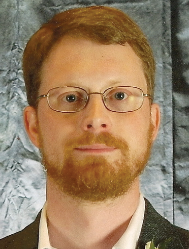Headshot of a man with red hair, wearing glasses and a suit