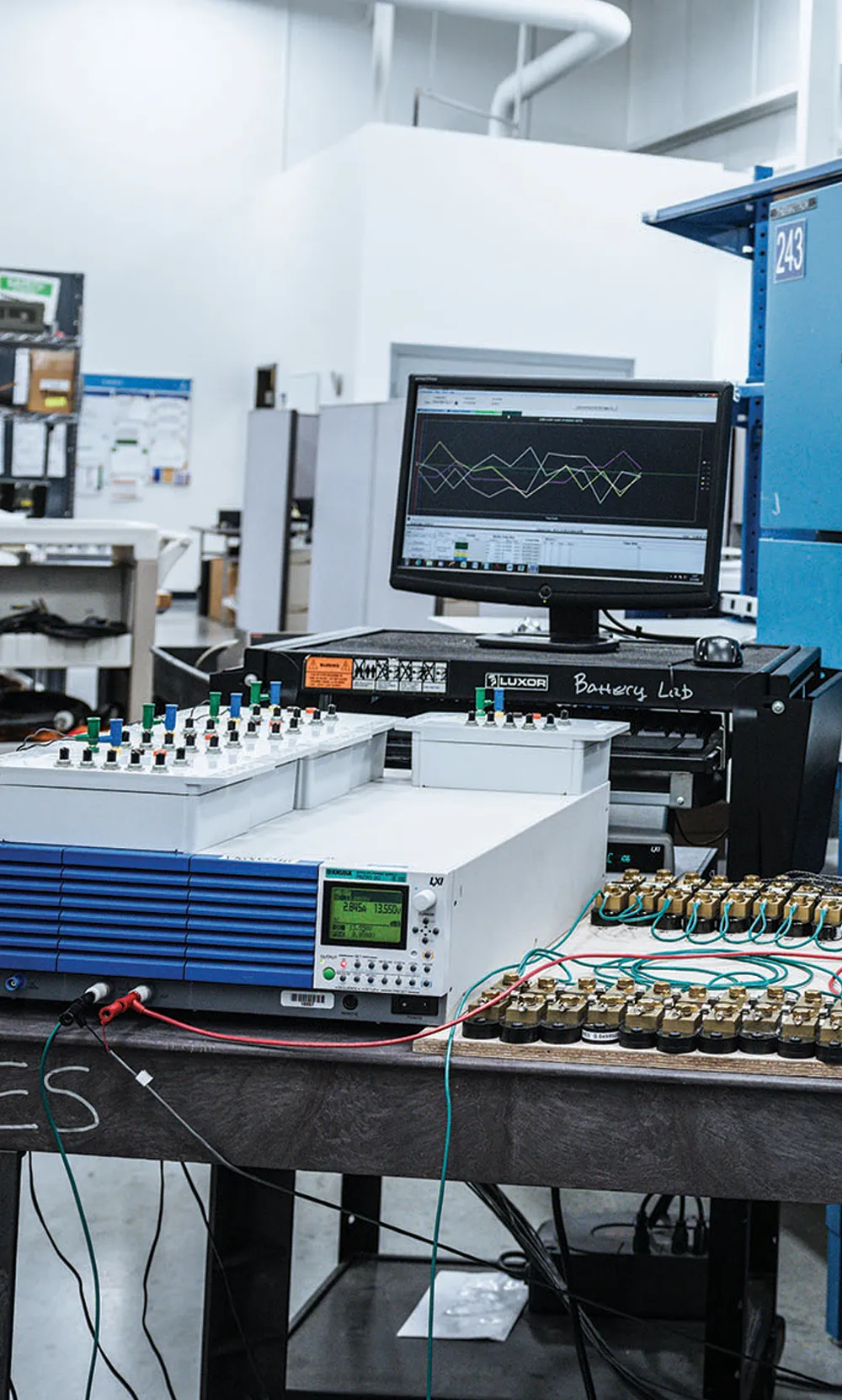 A laboratory setup with electronic testing equipment, including a computer monitor displaying data graphs, and a table with circuit boards and wires connected to testing devices. The environment appears to be industrial with various technical instruments organized on the workbench