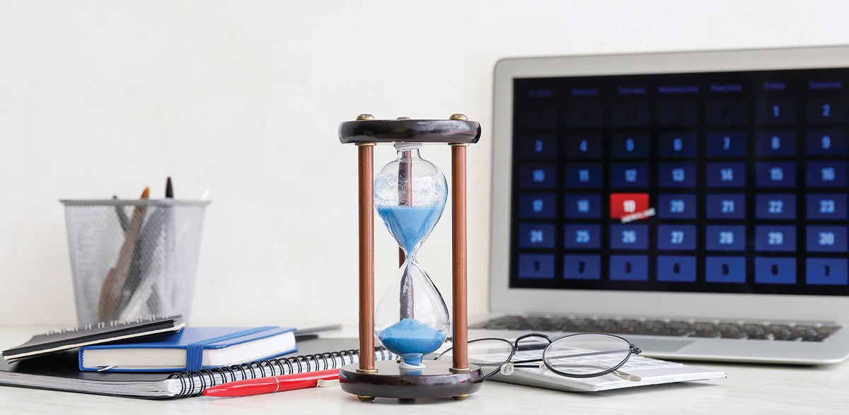 sandglass in front of a pile of notebooks, a cup of pens, eyeglasses, and a laptop