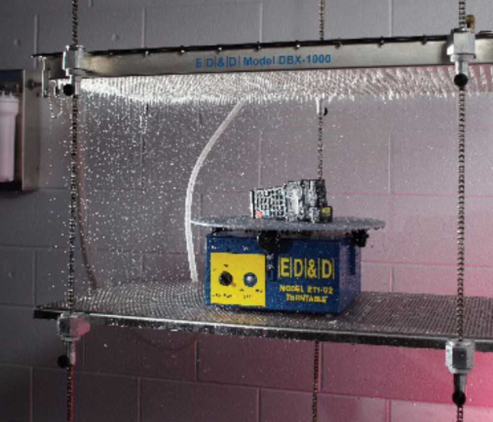 E D & D Model DBX-1000 water spray test apparatus showering a device placed on an E D & D Model 2T1-02 turntable, likely for environmental or ingress protection testing.