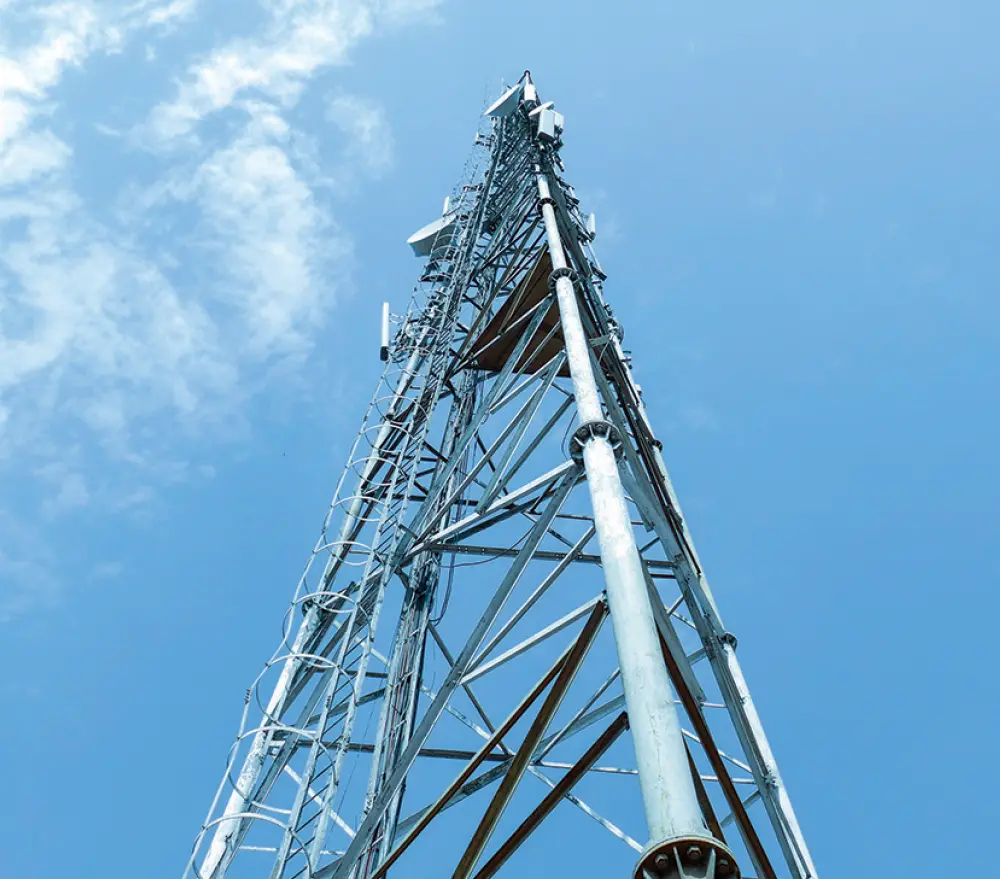A low-angle shot looking up at a tall, metallic cellular or radio tower structure against a clear blue sky with scattered white clouds; The tower's triangular lattice structure and antennas are visible