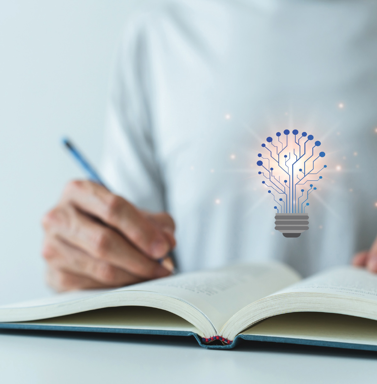 A person's hand holding a pen over an open book, with a glowing blue light bulb graphic made of circuit board lines floating above the pages.