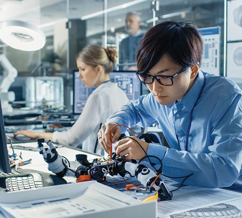 A male engineer in a blue shirt and glasses meticulously works on a robotic arm at a high-tech lab workbench, while a female colleague works in the background.