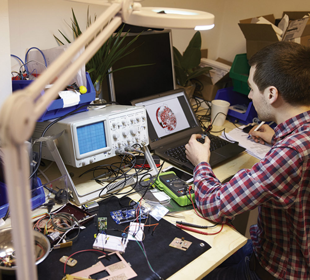 An engineer in a plaid shirt works at a cluttered electronics bench with an oscilloscope, a multimeter, and a laptop displaying a circular circuit board design.