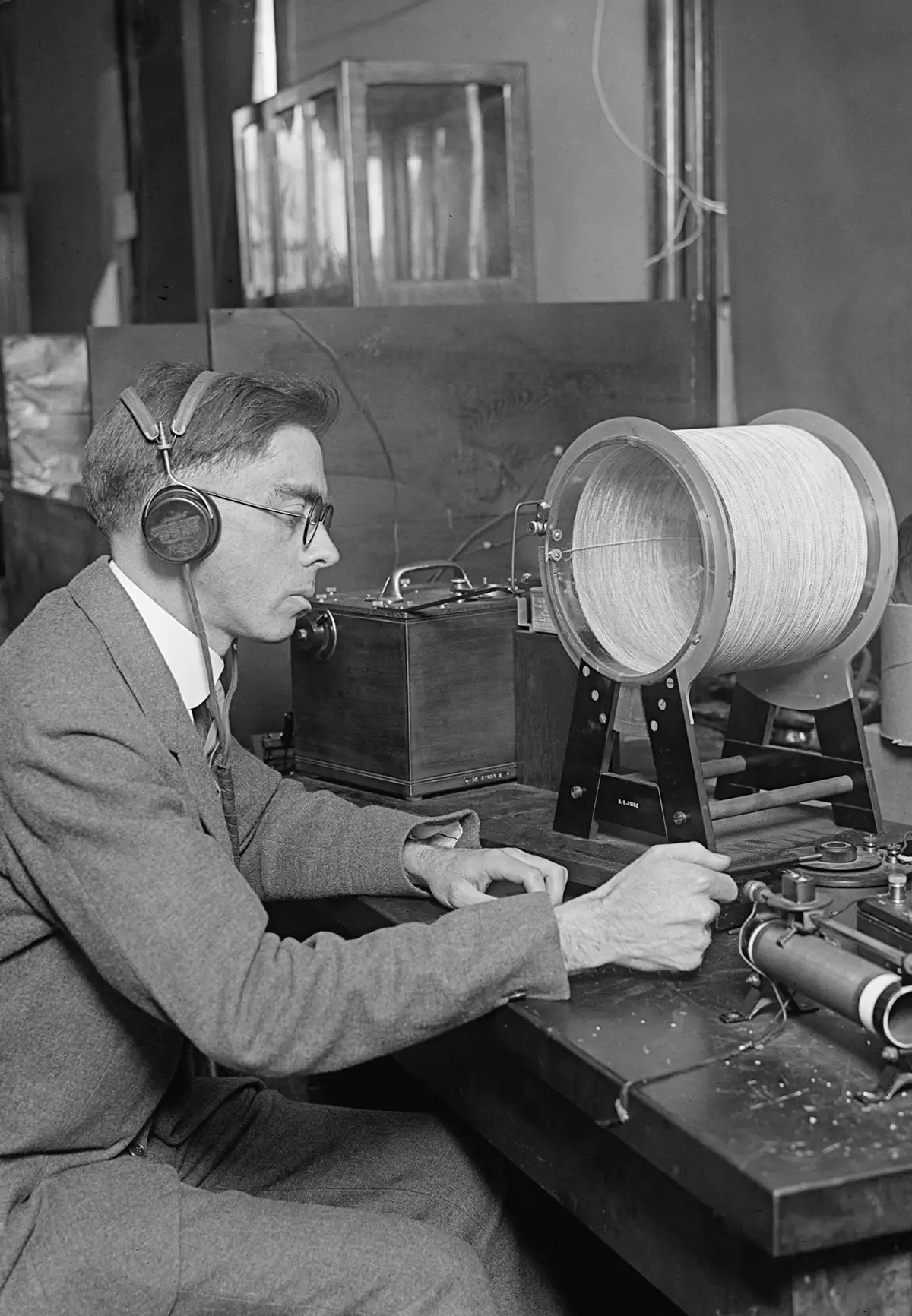 A vintage black-and-white photograph of a technician in a suit wearing headphones while operating a large, cylindrical early radio tuning coil on a wooden laboratory workbench
