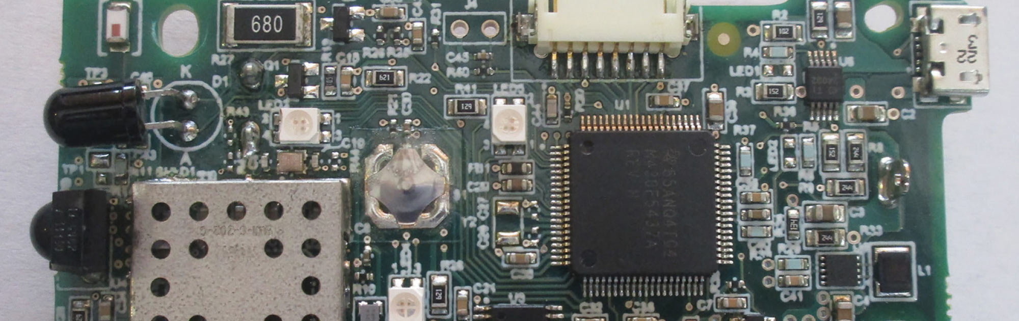 A close-up photograph of a green printed circuit board featuring a large central processor chip, various surface-mount components, and a metal shield.