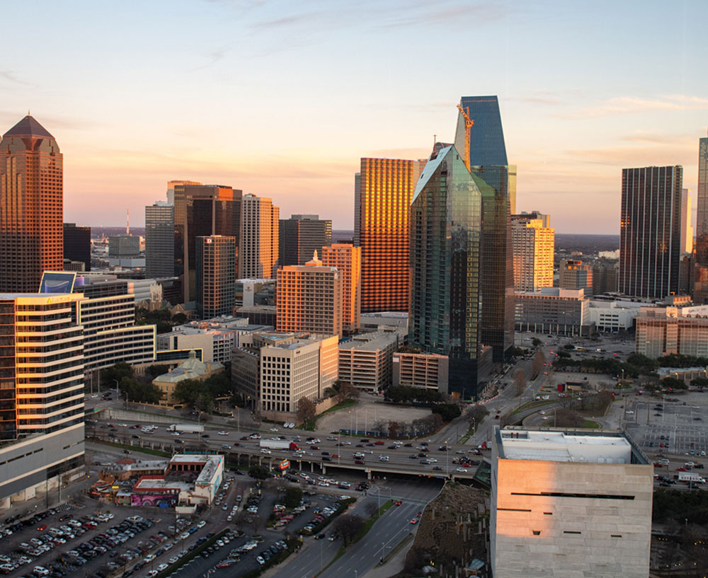Aerial view of the Dallas city skyline illuminated by a golden sunset, showing tall skyscrapers and highway traffic.