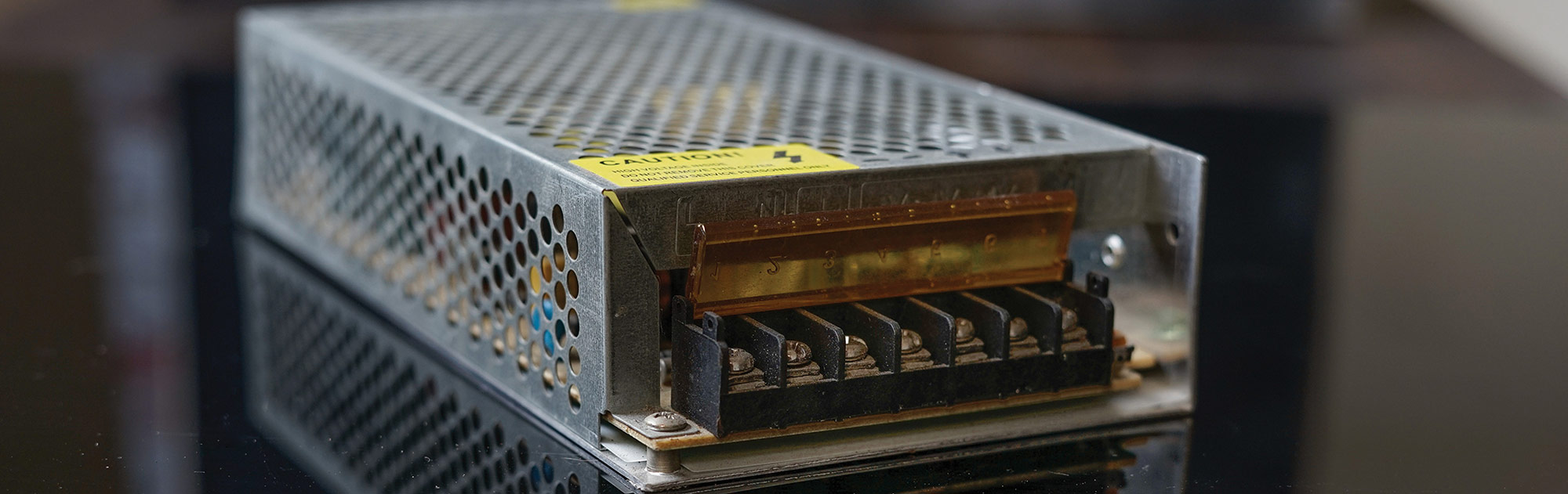A close-up of a metal-encased switching power supply featuring a perforated aluminum cover, a yellow caution sticker, and a screw terminal block under a translucent amber safety shield.