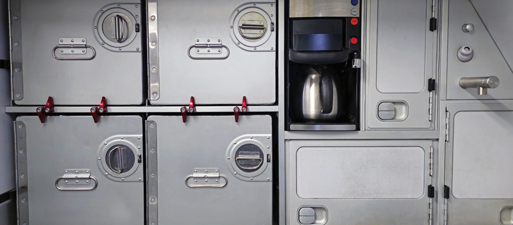 An airplane galley kitchen setup with metallic storage containers, red latches, and a coffee maker.