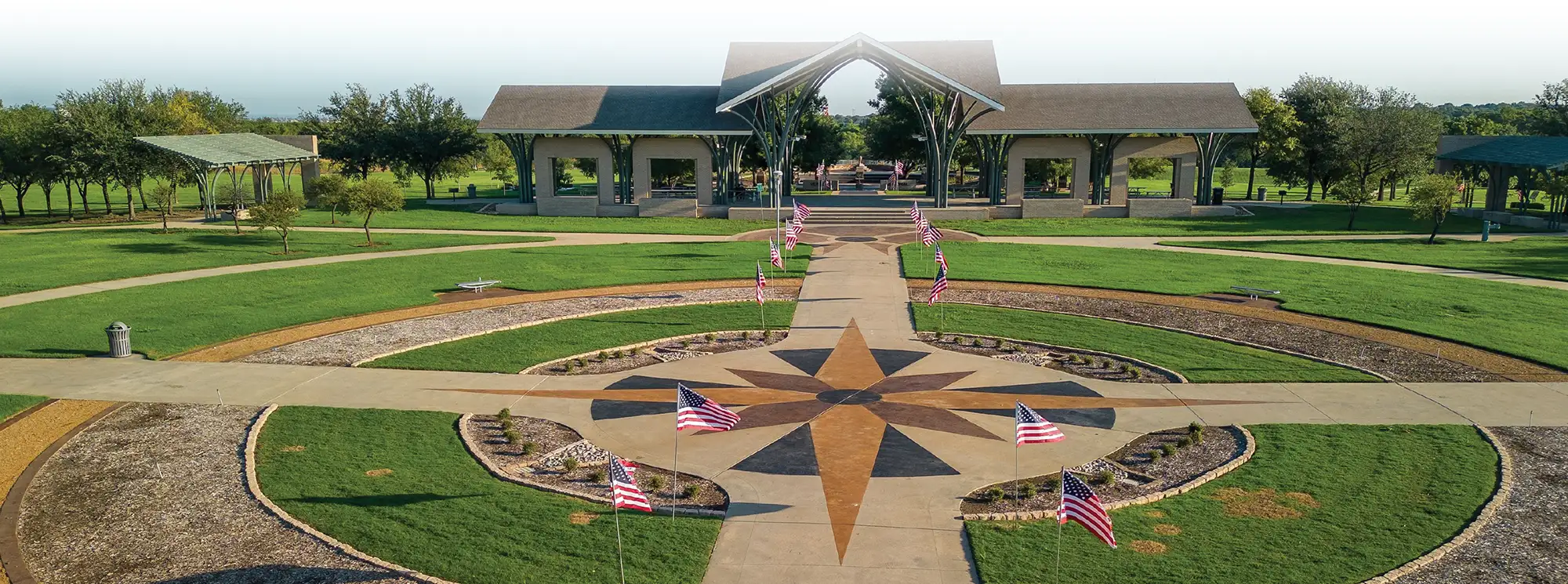Aerial view of a park plaza with a star pattern walkway, American flags, and a large pavilion.