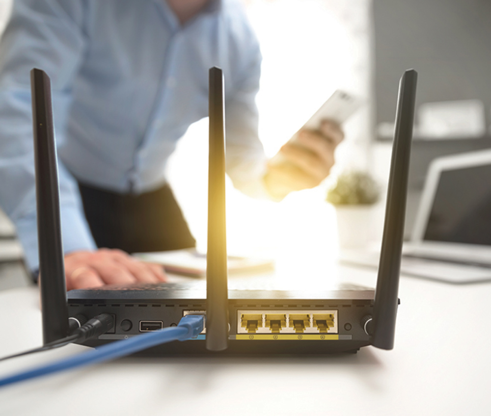 Close-up of a black Wi-Fi router with ethernet cables attached and a person using a smartphone.