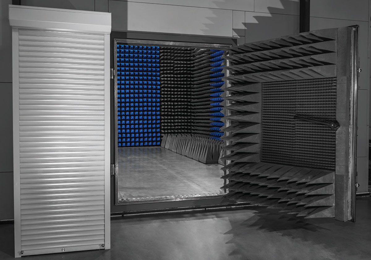 An open EMC anechoic test chamber showing an interior lined with blue and black RF absorber pyramids, with a heavy absorber-lined door open to the right and a white roll-up shutter to the left.