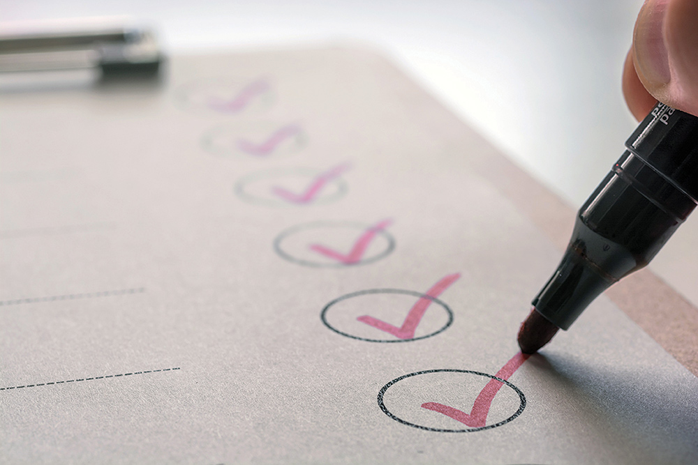A close-up view of a hand using a marker to mark pink checkmarks inside circled checkboxes on a paper checklist attached to a clipboard.